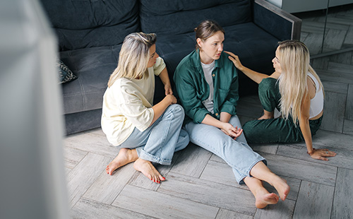 Drei Frauen sitzen vor einem Sofa auf dem Boden und reden miteinander. 